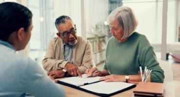 An older couple sitting at a table reviewing documents with a financial advisor. The man is pointing to a section on a paper while the woman holds a pen, listening attentively. They appear to be in a bright office or home setting with notebooks and pens on the table.