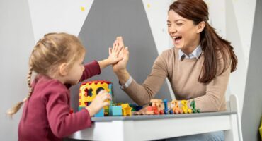 Adult and young child playing with colourful learning toys at a table and giving a high-five during an early childhood learning activity.