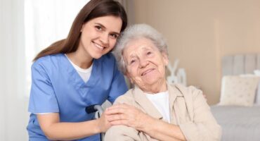 Smiling caregiver in blue scrubs supporting a happy elderly woman in a wheelchair at home.