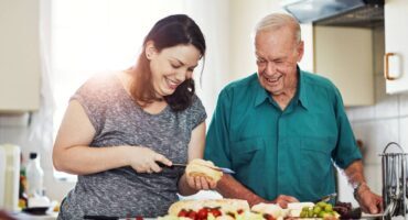Smiling caregiver and elderly man preparing a meal together in a bright kitchen, surrounded by fresh food like salad, grapes, and bread.