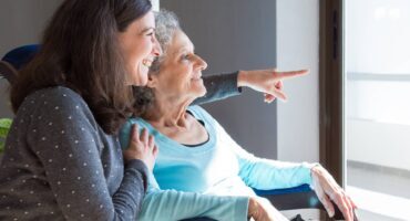 A smiling elderly woman in a wheelchair points out a window, sharing a joyful moment with a younger woman beside her.