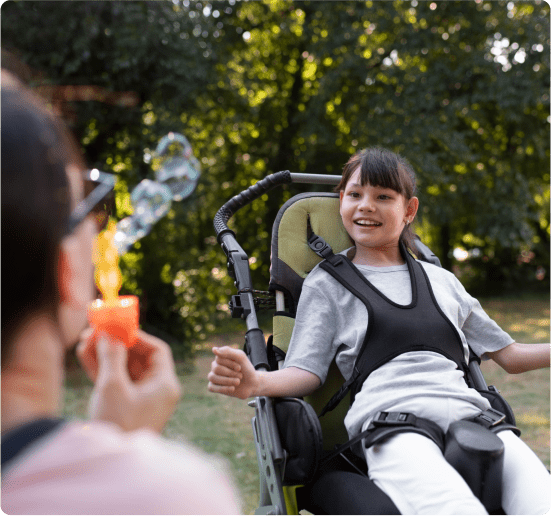 Smiling girl in a wheelchair watching an adult blow bubbles outdoors in a park