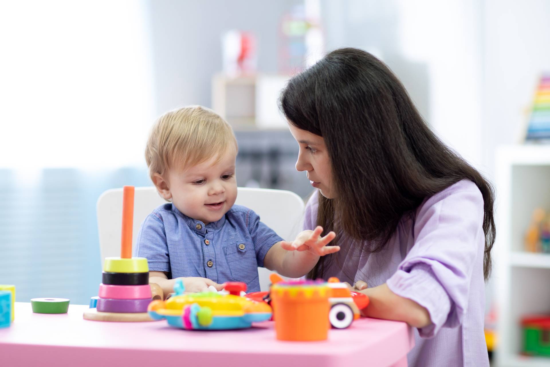 Woman engaging with a smiling toddler at a table filled with colorful toys in a bright playroom