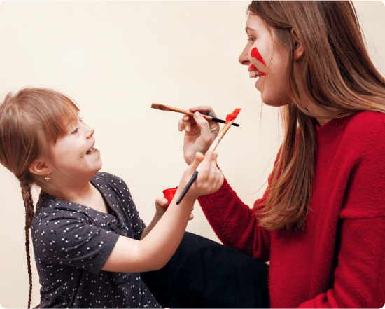 Young girl laughing while painting a woman's face with red paint, both smiling and having fun together
