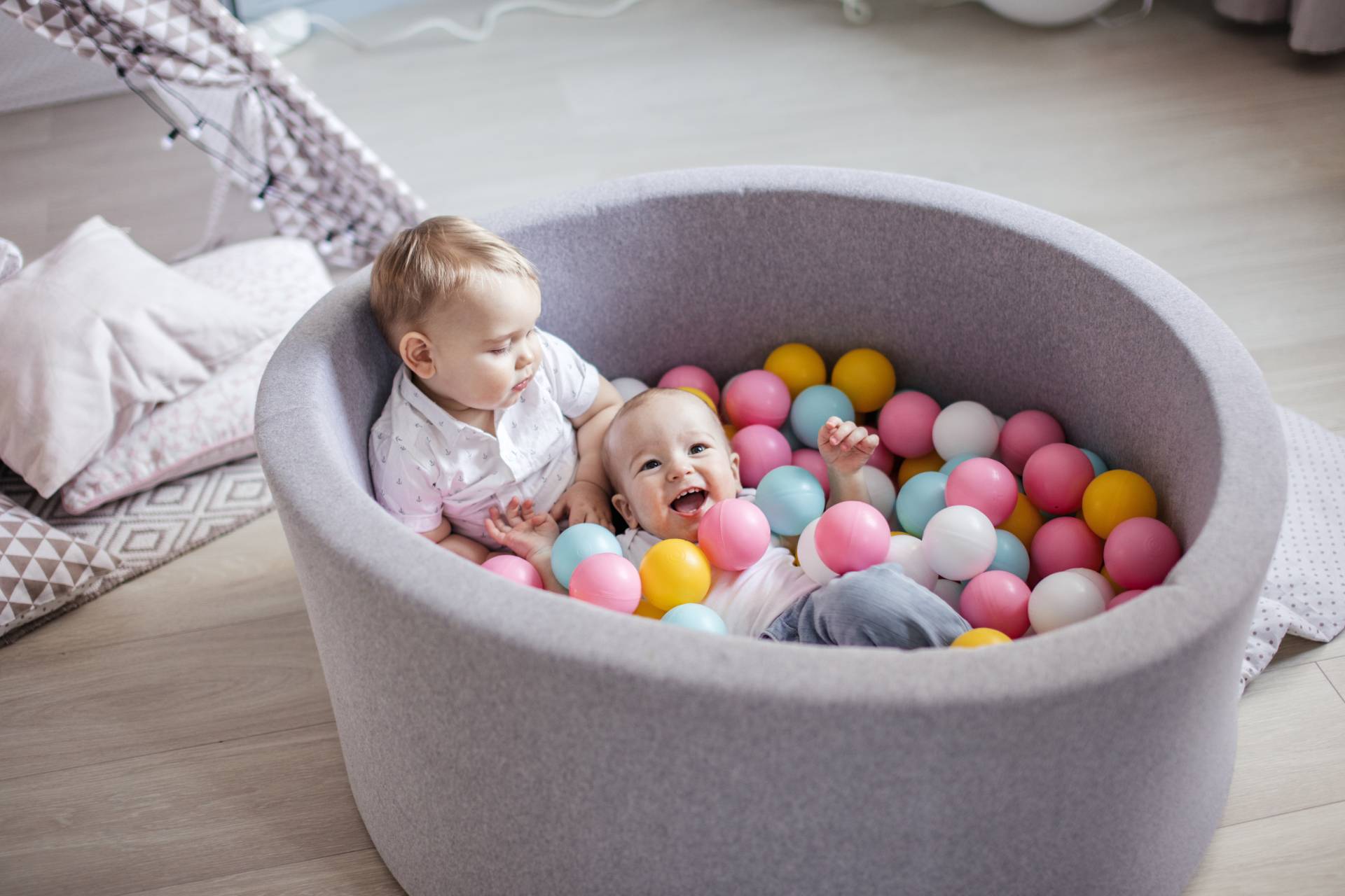 Two happy babies playing together in a gray felt ball pit filled with colorful pink, yellow, blue, and white balls