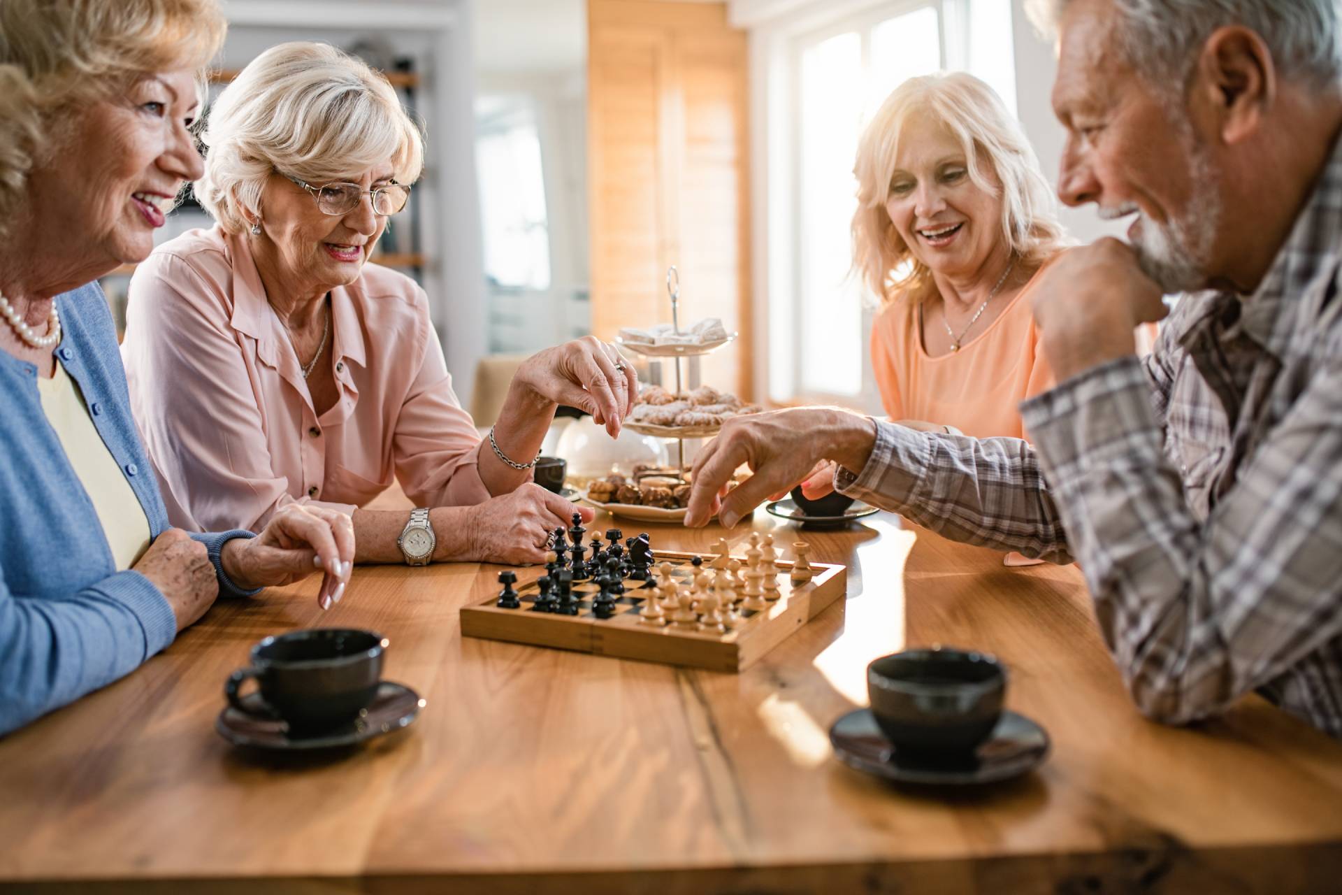 Two seniors have fun playing chess while friends watch and drink coffee