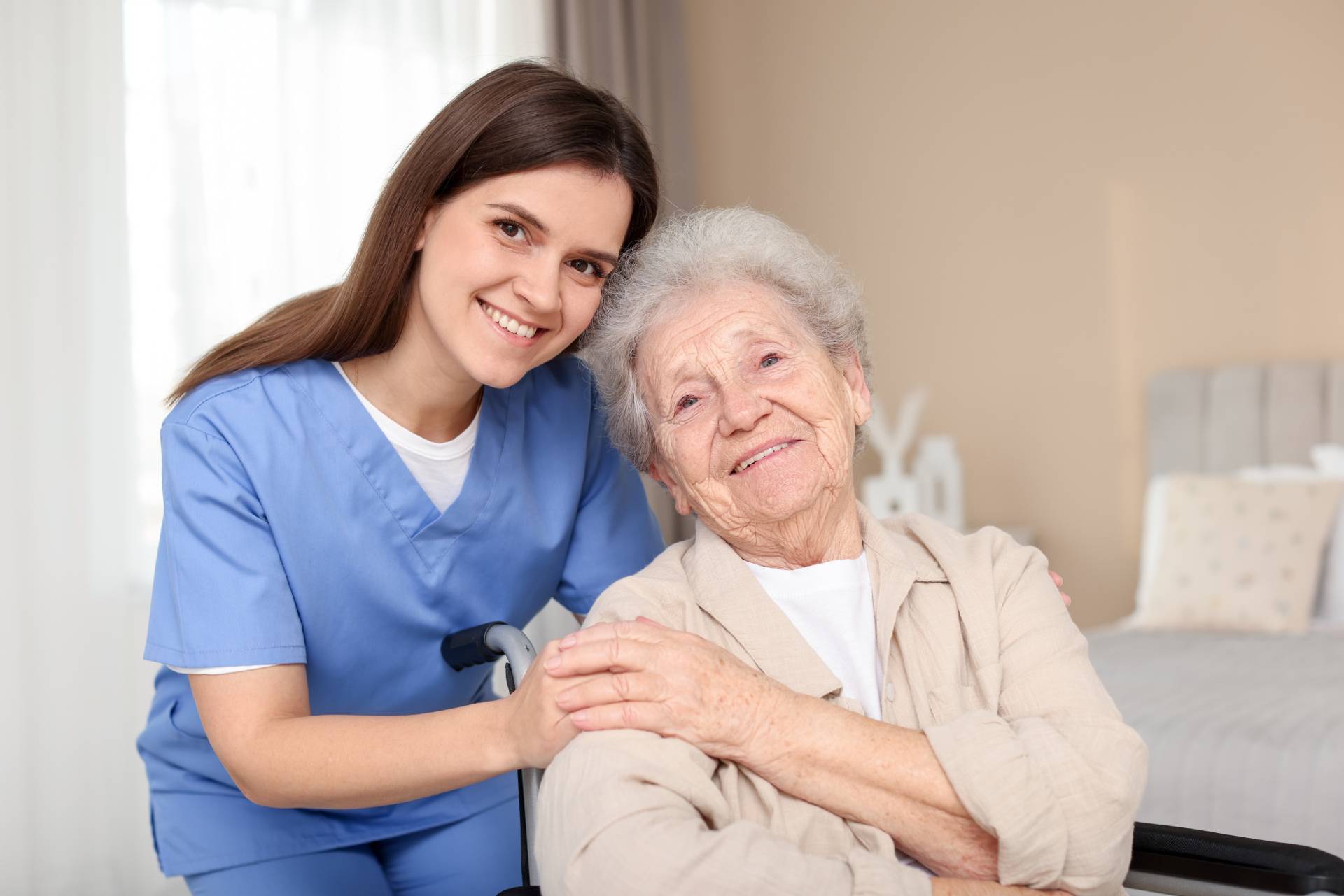 Smiling caregiver in blue scrubs supporting a happy elderly woman in a wheelchair at home.