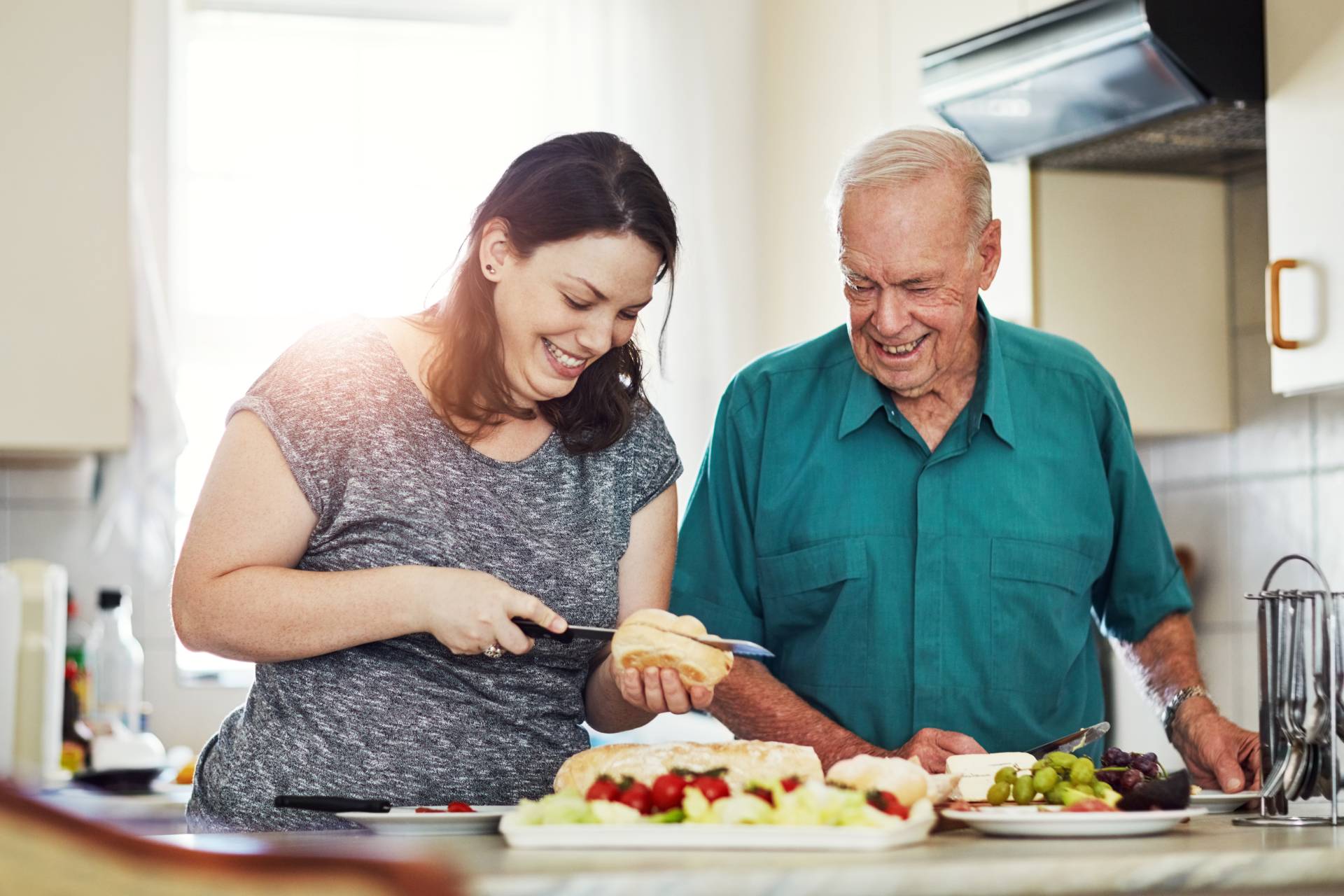 Smiling caregiver and elderly man preparing a meal together in a bright kitchen, surrounded by fresh food like salad, grapes, and bread.