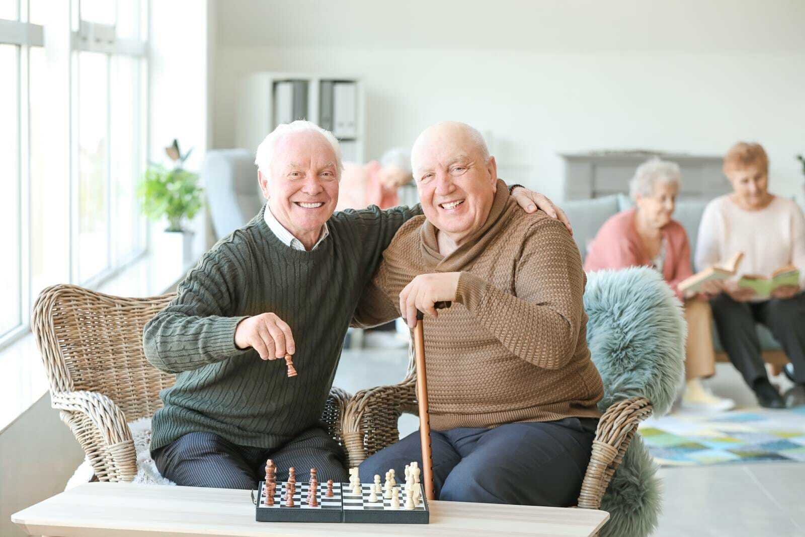 Two men with dementia with their arms around one another in front of a chess board.