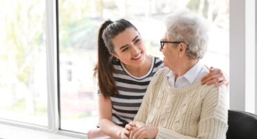 Caregiver with senior woman in nursing home A young woman smiles warmly at an elderly woman, gently holding her hands and sitting close together by a large bright window.