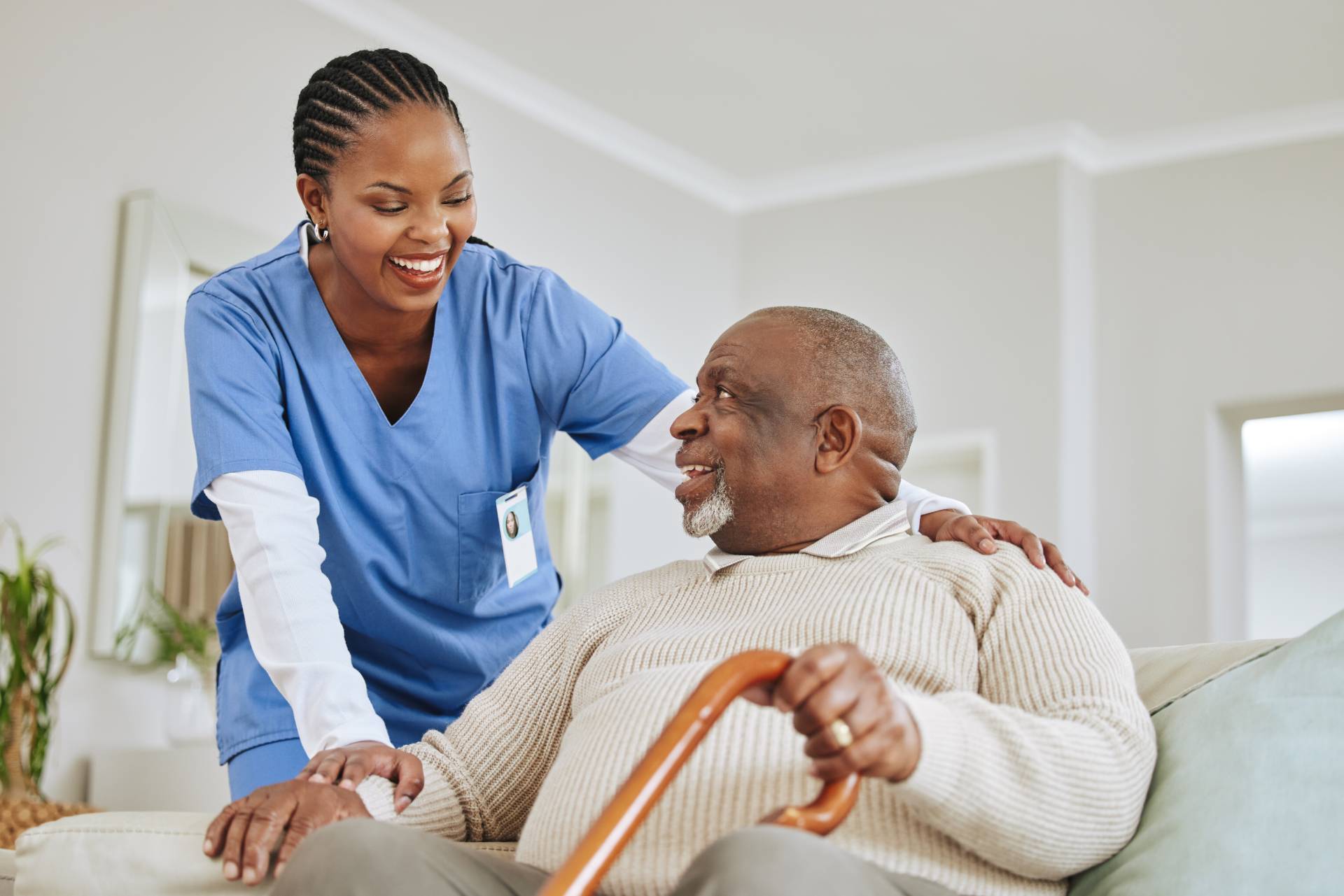 Smiling caregiver in blue scrubs supporting an elderly man with a cane at home