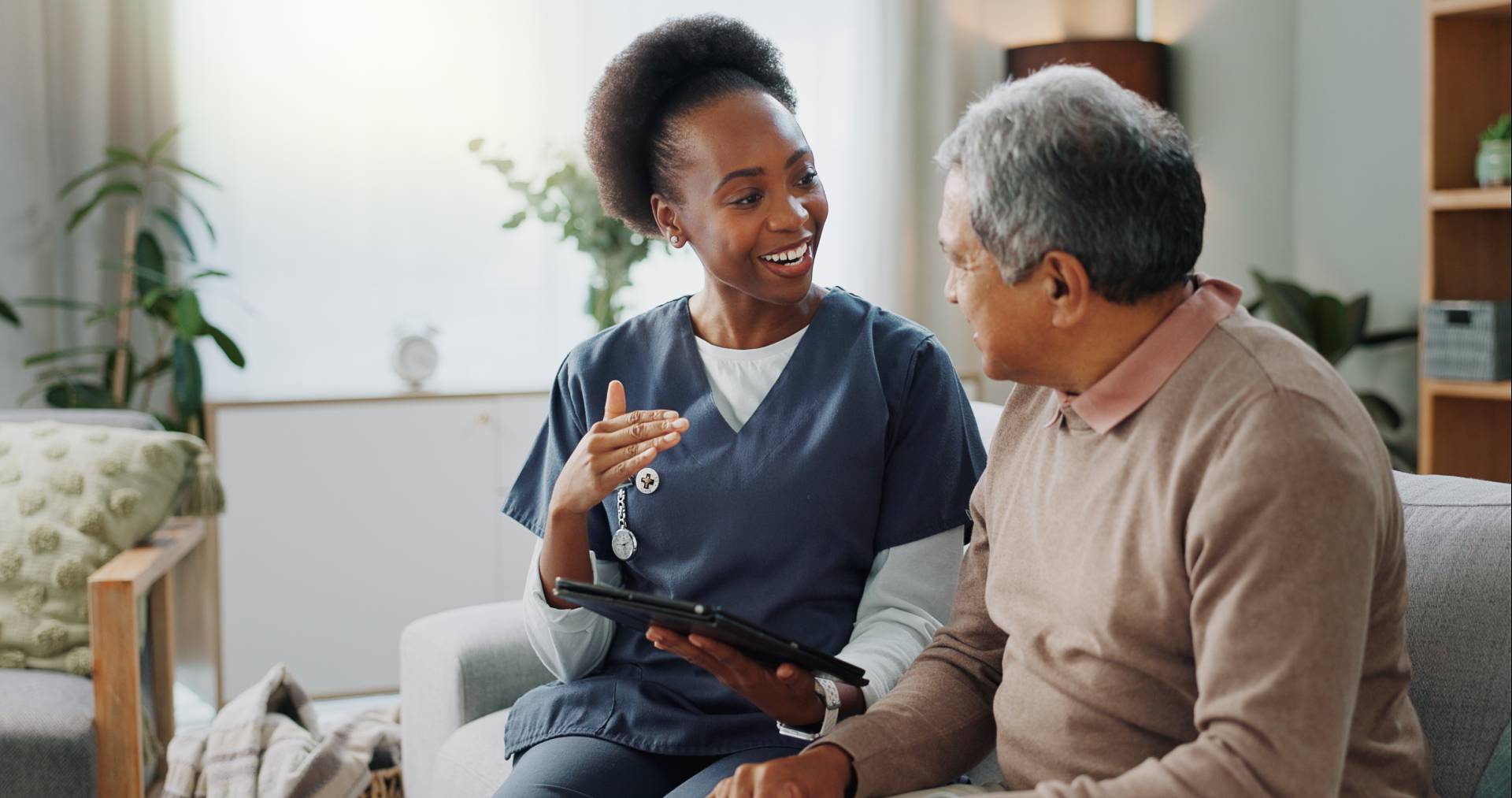 A caregiver in navy scrubs sits on a couch with an older man, smiling and gesturing while holding a tablet as they talk in a bright, comfortable living room.