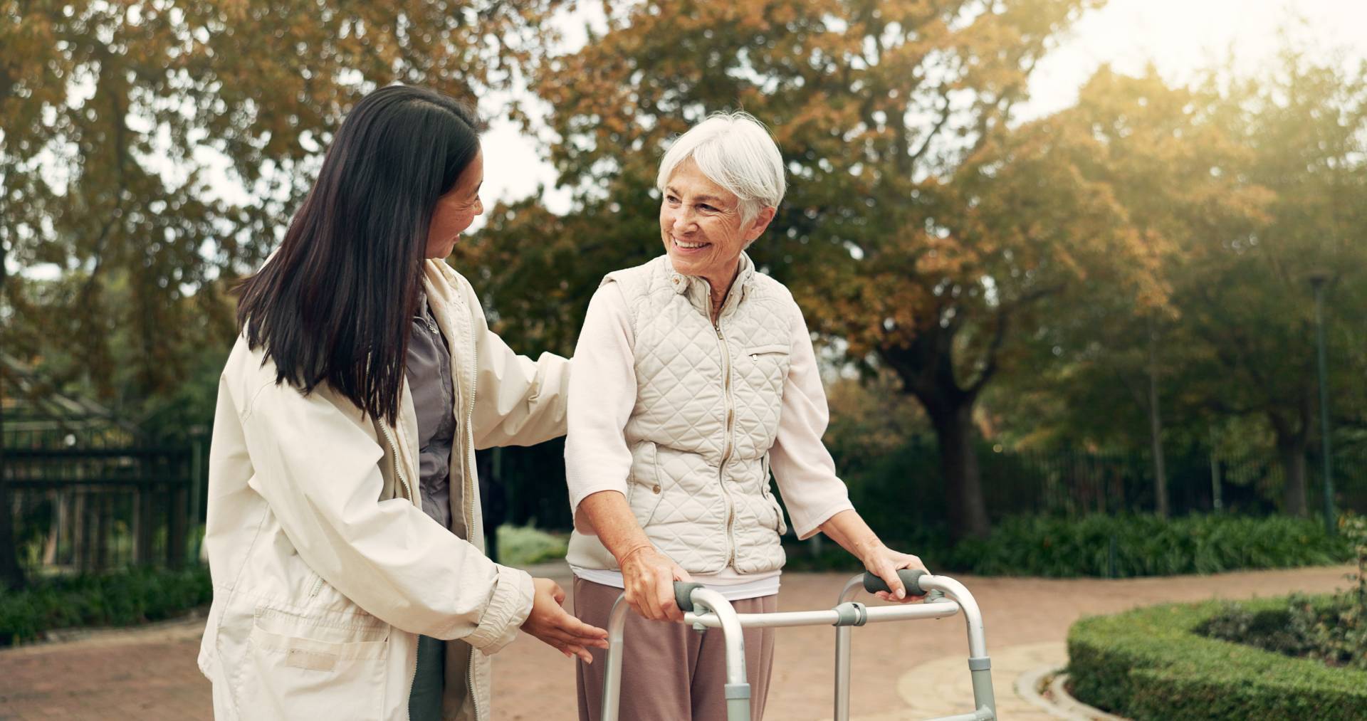 Caregiver supporting an elderly woman using a walker while walking outdoors in a park, both smiling and enjoying the moment.