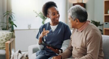 advice-for-homecare A caregiver in navy scrubs sits on a couch with an older man, smiling and gesturing while holding a tablet as they talk in a bright, comfortable living room.