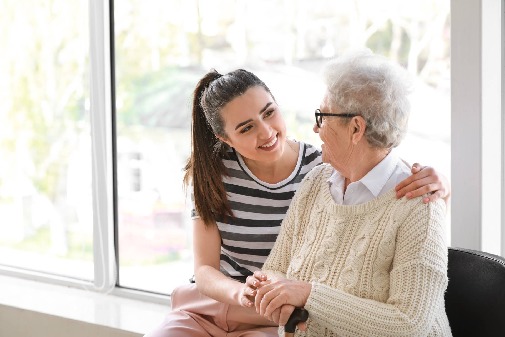 A young woman smiles warmly at an elderly woman, gently holding her hands and sitting close together by a large bright window.