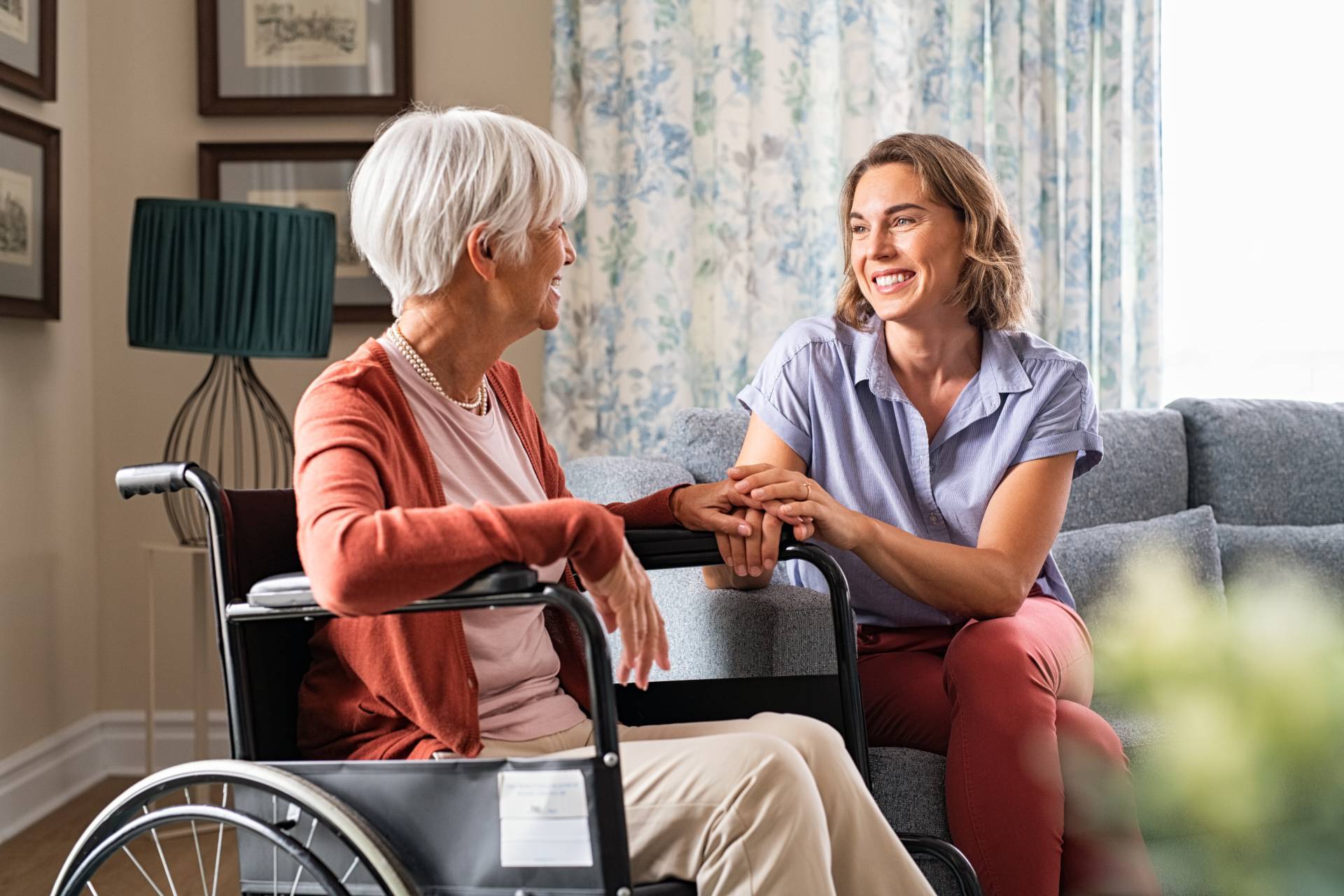Smiling woman sitting on a couch holding hands with an elderly woman in a wheelchair at home.