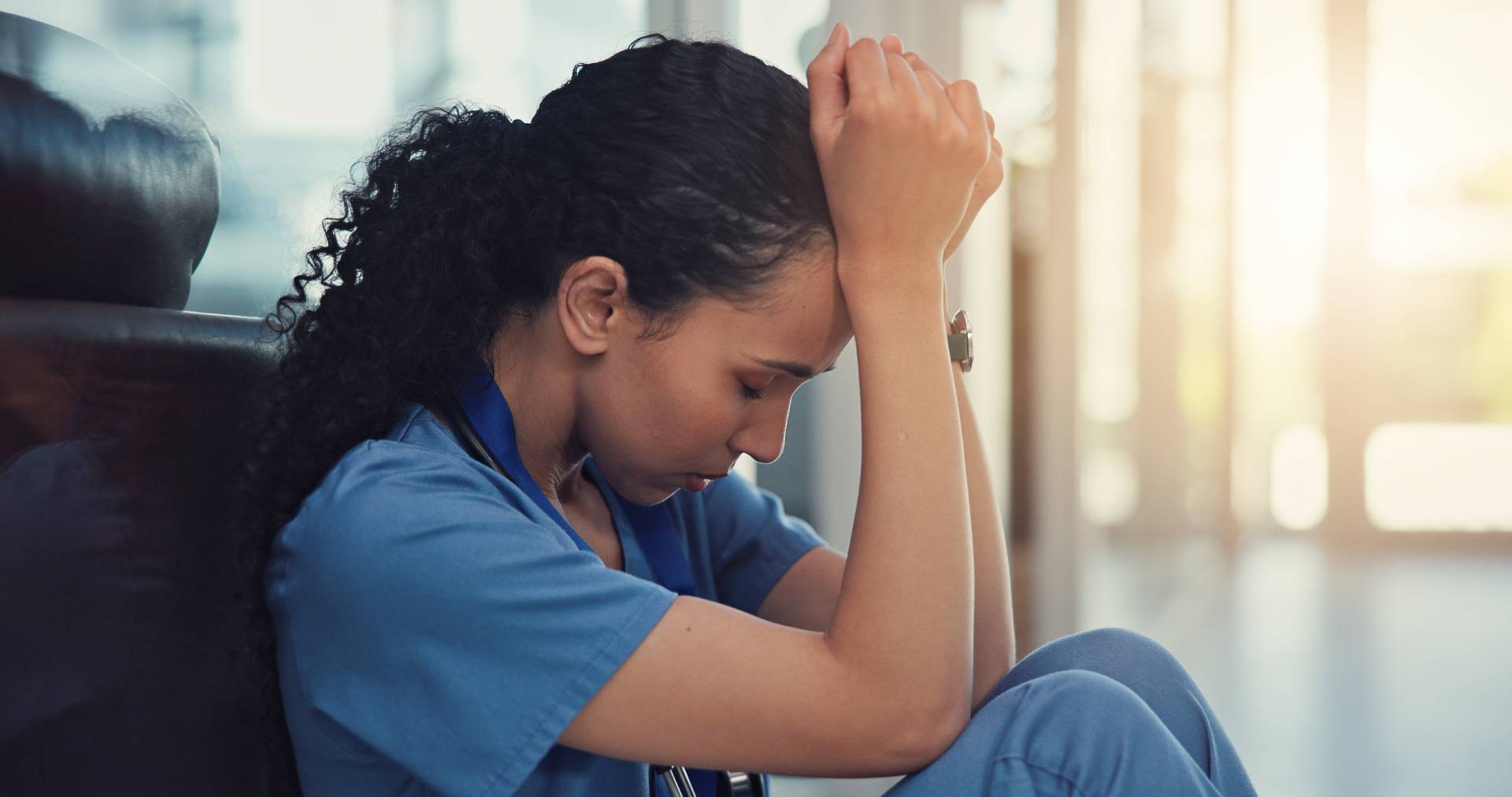 Exhausted healthcare worker in scrubs sitting on the floor with head bowed and hands clasped, showing signs of stress and fatigue.