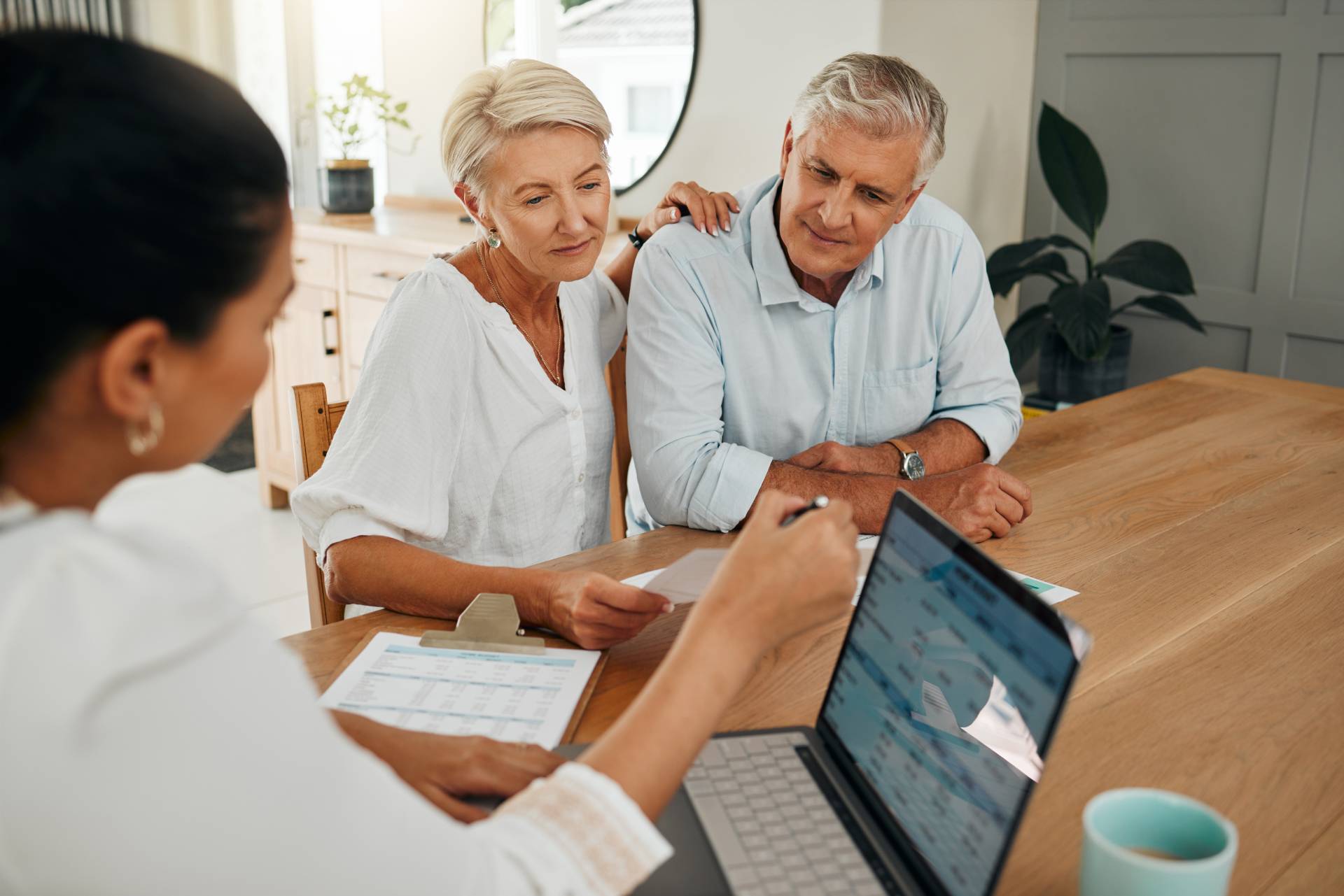An older couple sitting at a wooden table attentively listening to a financial advisor who is showing information on a laptop. The woman has her hand on the man’s shoulder, and documents with charts and a clipboard are visible on the table in a bright, modern home setting.