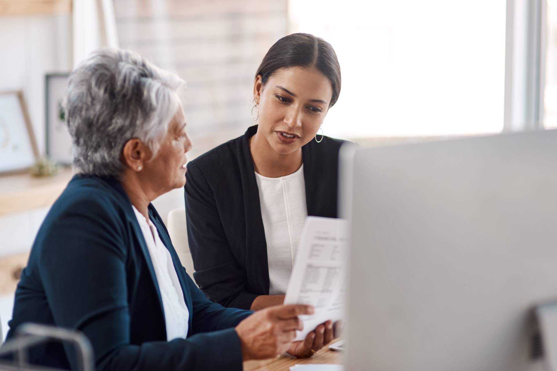 A younger woman in professional attire reviewing a document with an older senior woman at a desk, discussing care options and costs.