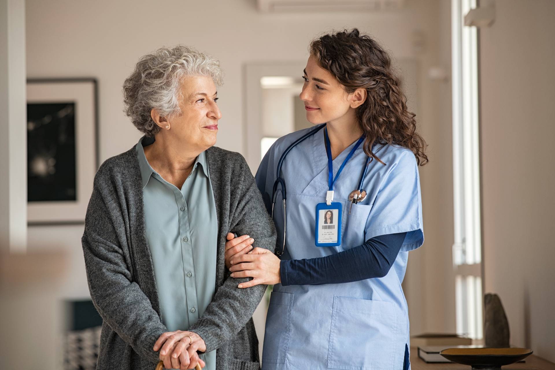 Female nurse in scrubs supporting and smiling at an elderly woman with a cane