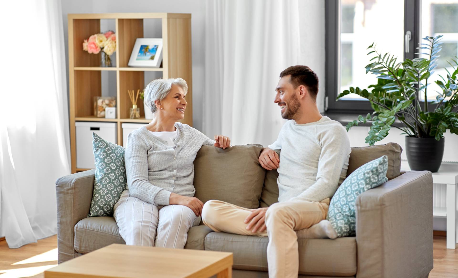 Smiling senior woman and adult man sitting on a couch and chatting in a cozy, modern living room with decorative pillows and houseplants.