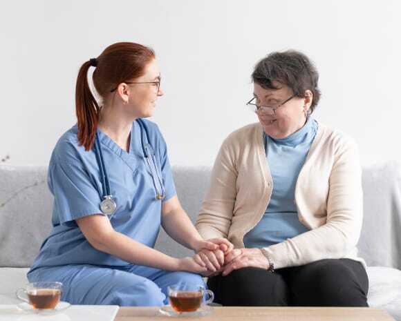 A nurse in blue scrubs and glasses gently holds hands with an elderly woman in a beige cardigan. Both are smiling, seated on a couch with tea cups in front.