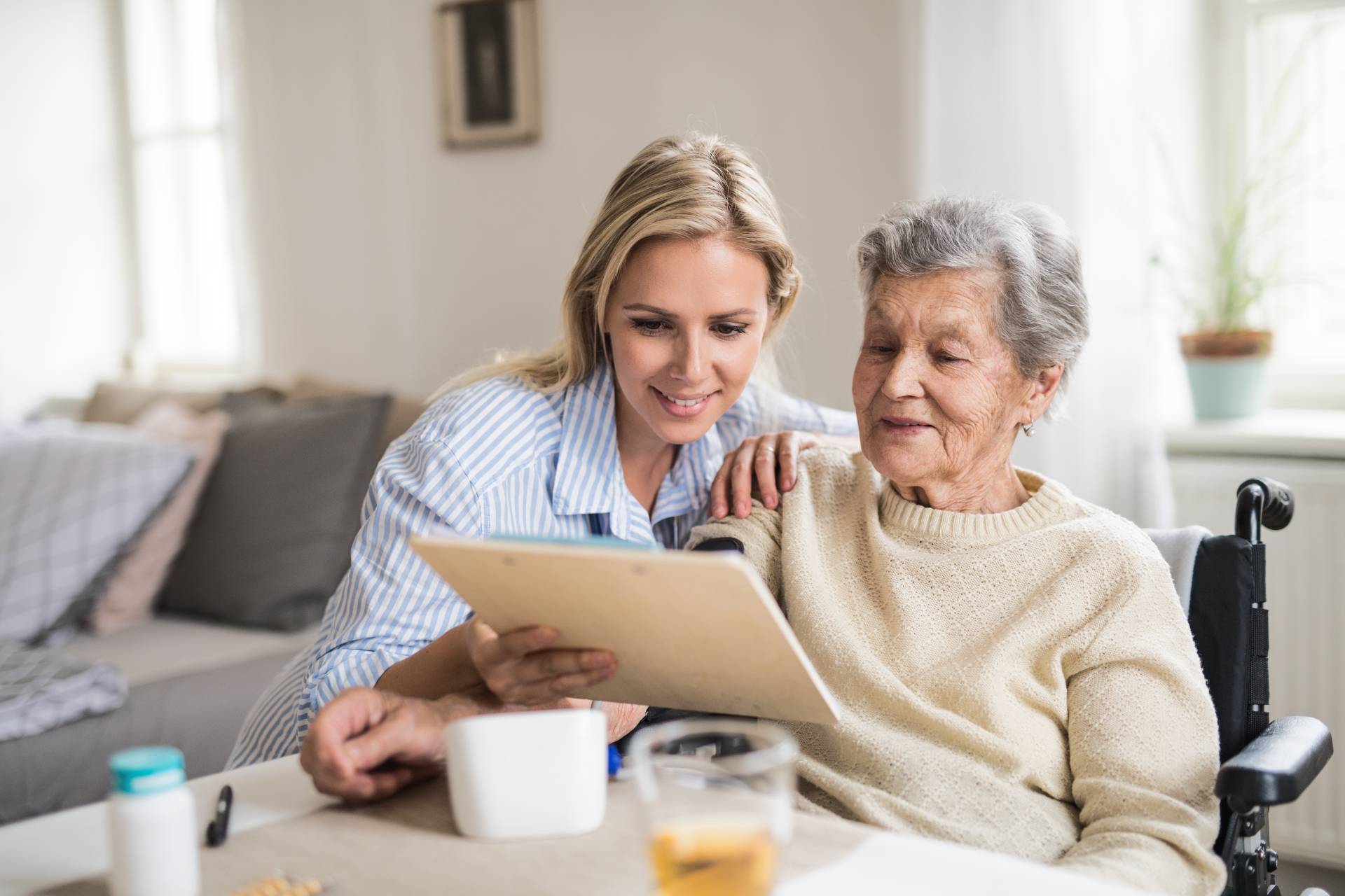 A younger woman sits beside an older woman in a wheelchair, smiling as they look together at a tablet in a bright, cozy living room.