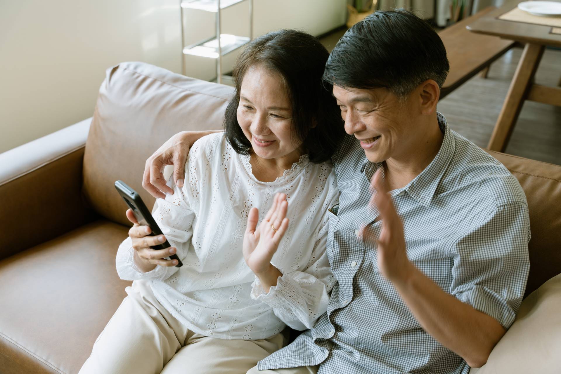 Two seniors sitting on a couch, waving at their phone as they are on a video call with a family member.