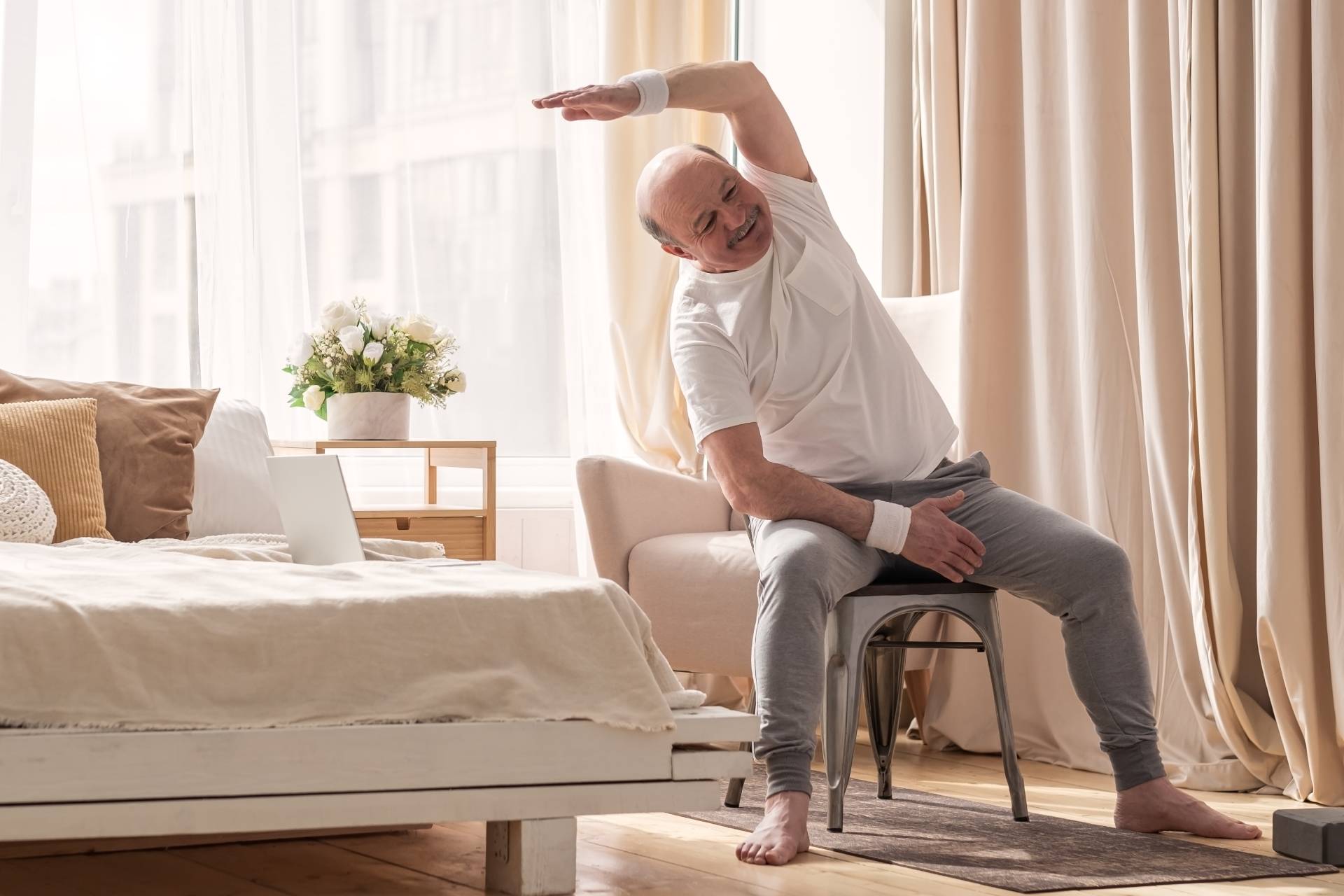 "Senior man doing arm chair yoga with a side stretch in a cozy bedroom, demonstrating gentle seated yoga for seniors."