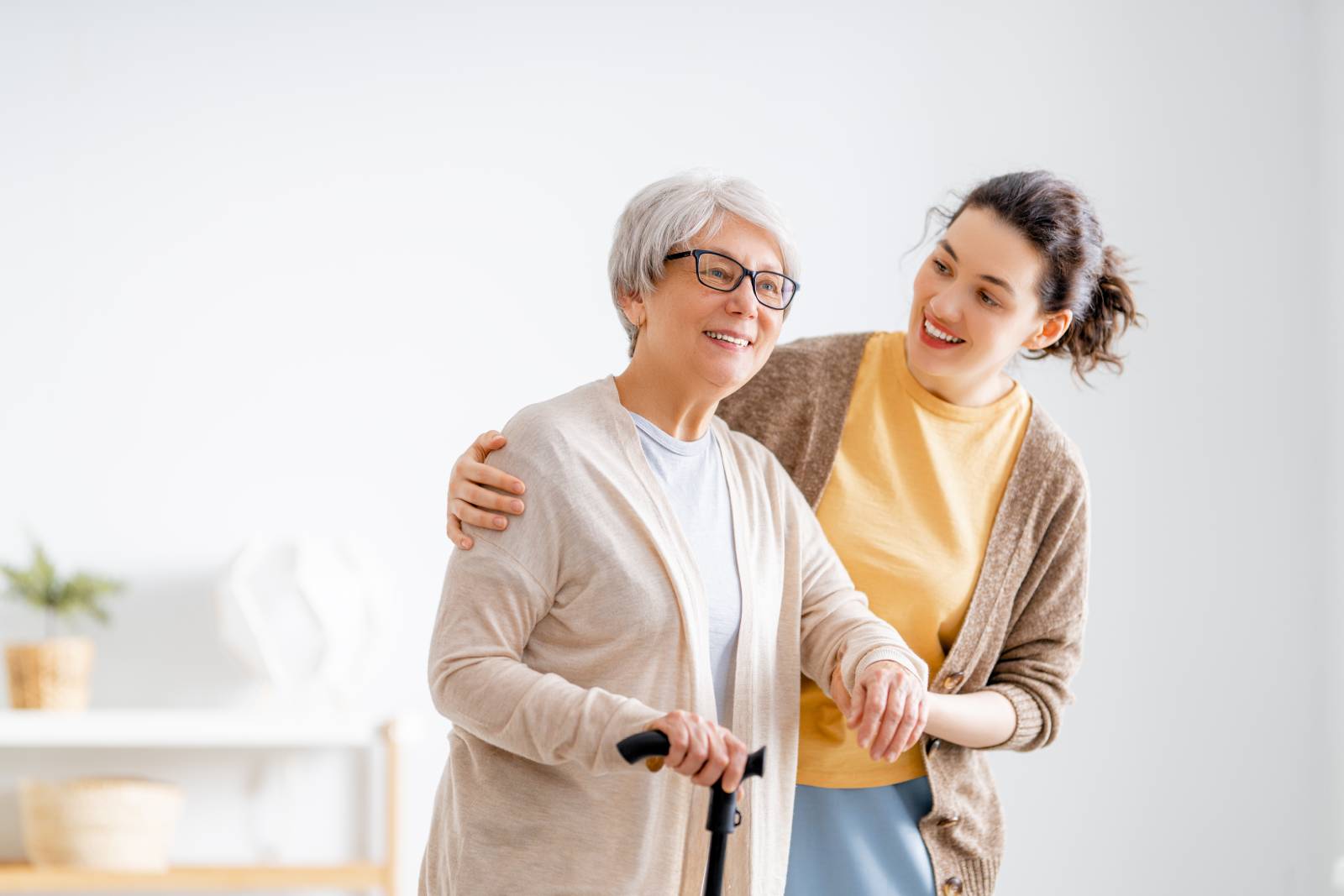 A young woman supports an elderly woman with short white hair using a cane. They are both smiling as they look ahead in a bright, modern living room