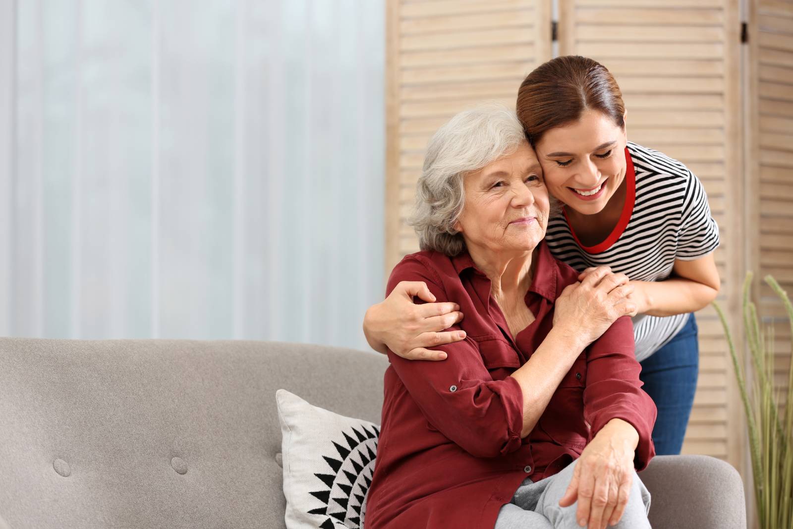 A smiling young woman embracing an elderly woman who is sitting on a couch. The elderly woman appears content and is looking affectionately at the younger woman. They are in a bright, cozy room, suggesting a close and caring relationship, possibly between a caregiver and a senior family member.