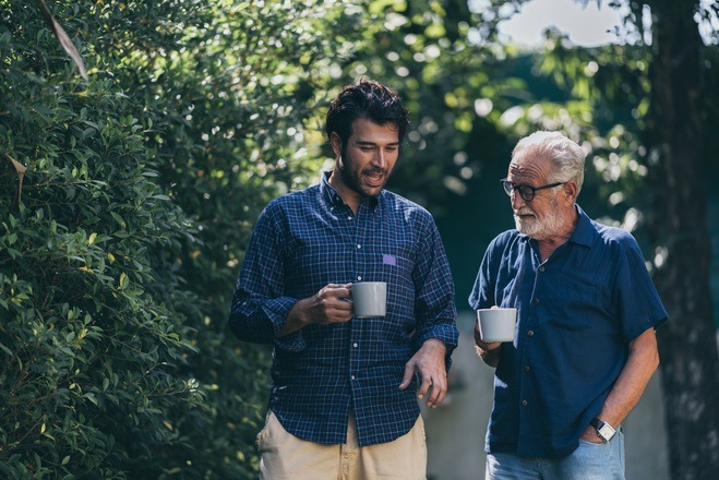 A young man and an elderly man walk through a lush garden, both holding white coffee mugs. The young man, wearing a blue and black checkered shirt, smiles and talks animatedly, while the elder, sporting a blue shirt and glasses, listens attentively with a slight smile. The background is filled with dense green foliage