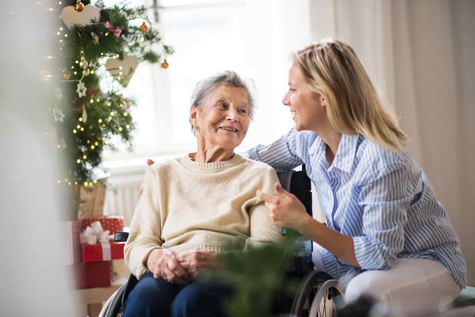 n older woman in a wheelchair smiling warmly at a younger woman beside her, who is talking and gesturing with a cheerful expression. They are sitting near a decorated Christmas tree with wrapped presents underneath, in a cozy, sunlit room.