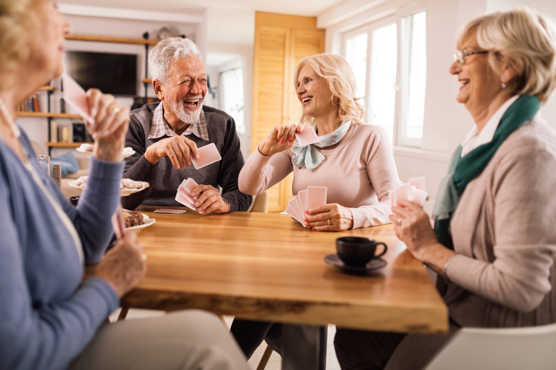 A group of older adults laughing and playing cards around a table with coffee and snacks.