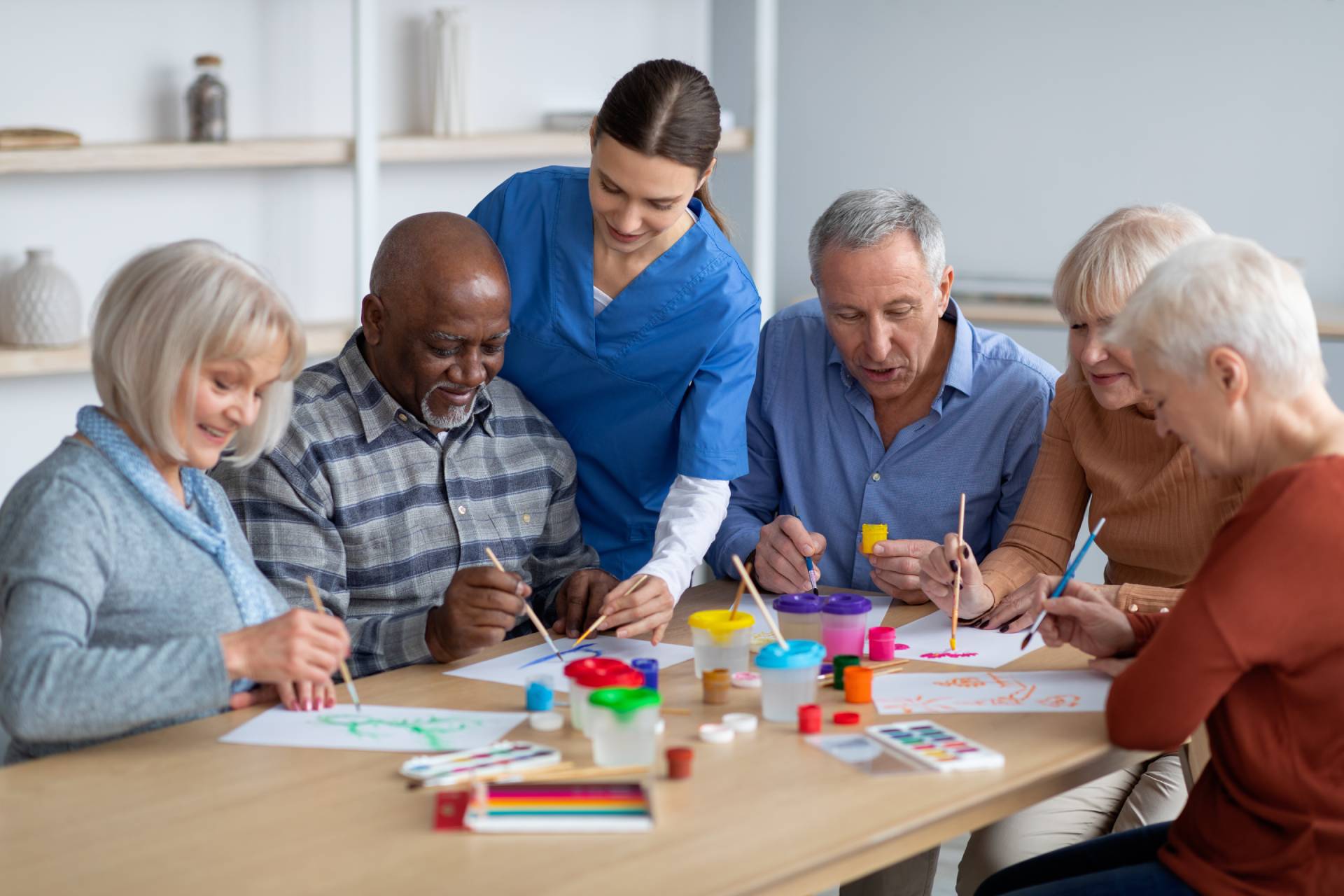 Caregiver helps a group of older adults paint together at a table with art supplies.