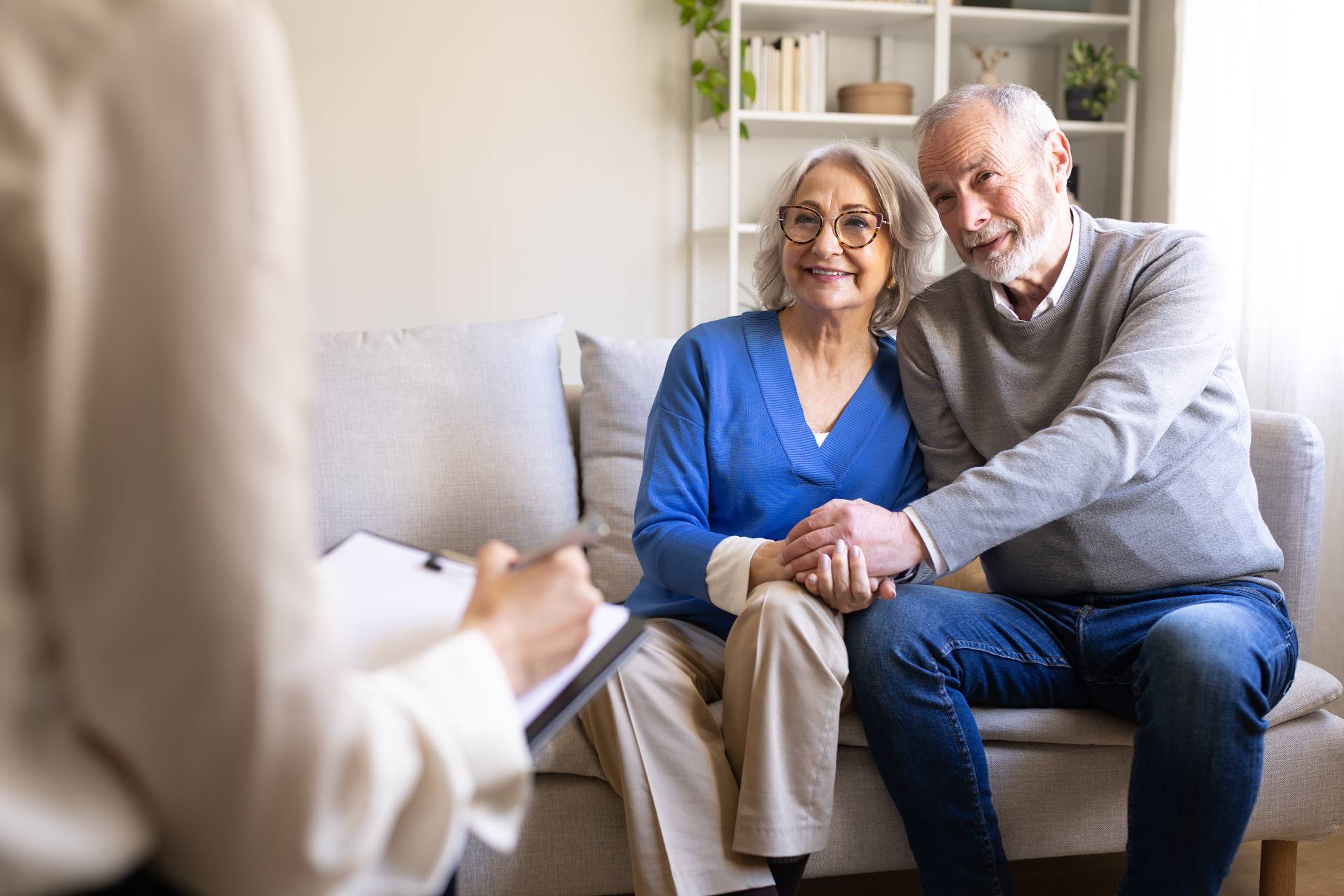A senior couple sitting together on a couch holding hands and smiling while meeting with a care advisor taking notes on a clipboard.