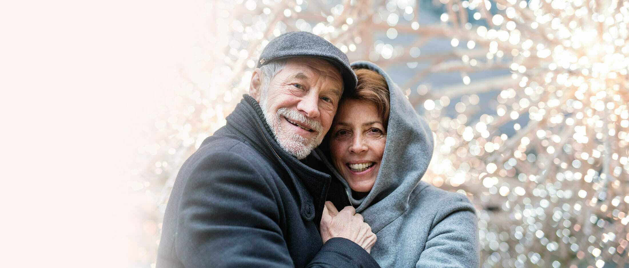An elderly man and woman hugging in front of Christmas lights.
