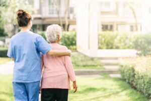 A caregiver in blue scrubs walking outdoors with an elderly woman in a garden setting.