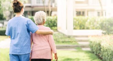 A caregiver in blue scrubs walking outdoors with an elderly woman in a garden setting.