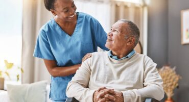home-care-comparison Smiling home care nurse in blue scrubs standing beside an elderly man in a wheelchair, gently resting her hand on his shoulder as they look at each other in a bright living room.