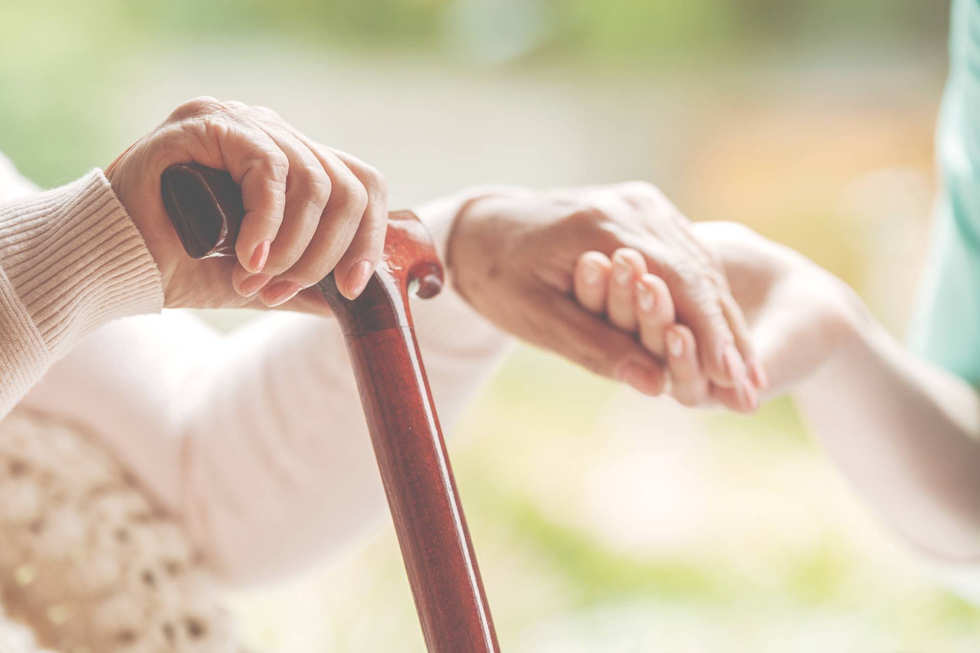 A close-up of an older adult’s hand holding a wooden cane while a caregiver gently rests their hand on top, offering support.