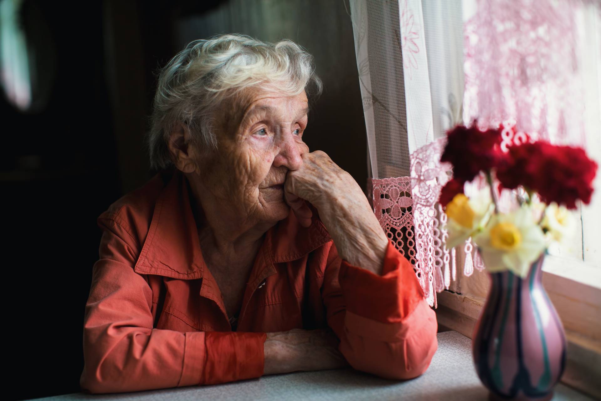 Elderly woman with gray hair, resting her chin on her hand as she looks pensively out of a window, with a vase of flowers on the table beside her.