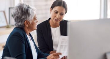 A younger woman in professional attire reviewing a document with an older senior woman at a desk, discussing care options and costs.