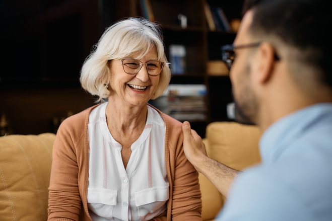 A joyful elderly woman with white hair and glasses shares a laugh in a warm, light-filled room. She wears a peach cardigan over a white blouse and is engaging in a pleasant conversation with a man, visible from the back, who is gesturing while wearing a blue shirt.