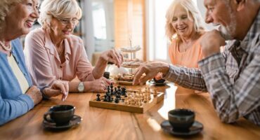 Two seniors have fun playing chess while friends watch and drink coffee