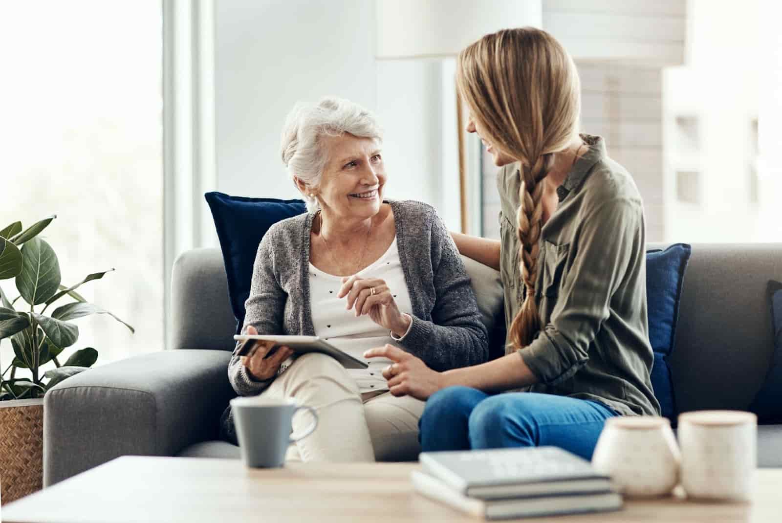 n older woman and a younger woman sitting together on a couch, smiling and talking while looking at a tablet. The setting appears to be a bright, cozy living room with natural light, blue pillows, and a coffee table with mugs and books.