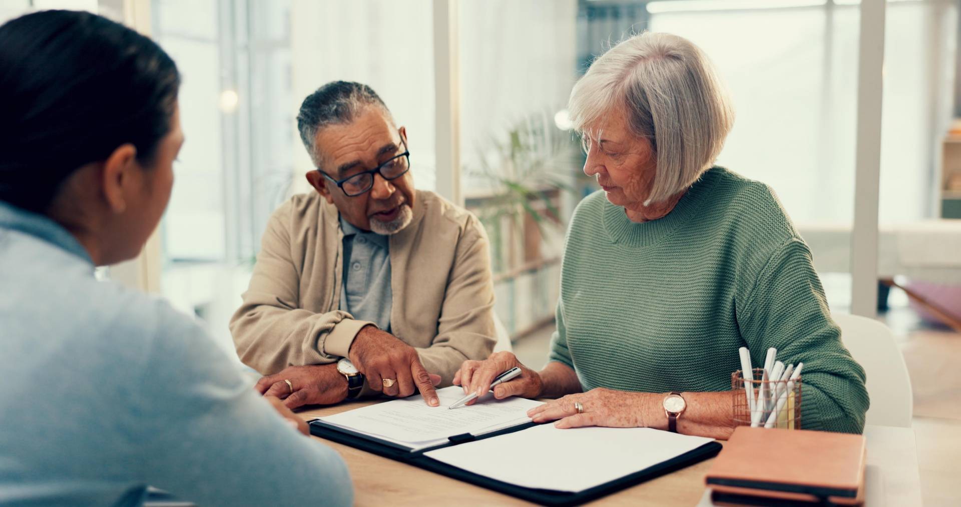 An older couple sitting at a table reviewing documents with a financial advisor. The man is pointing to a section on a paper while the woman holds a pen, listening attentively. They appear to be in a bright office or home setting with notebooks and pens on the table.