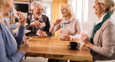 A group of older adults laughing and playing cards around a table with coffee and snacks.