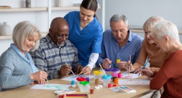 Caregiver helps a group of older adults paint together at a table with art supplies.