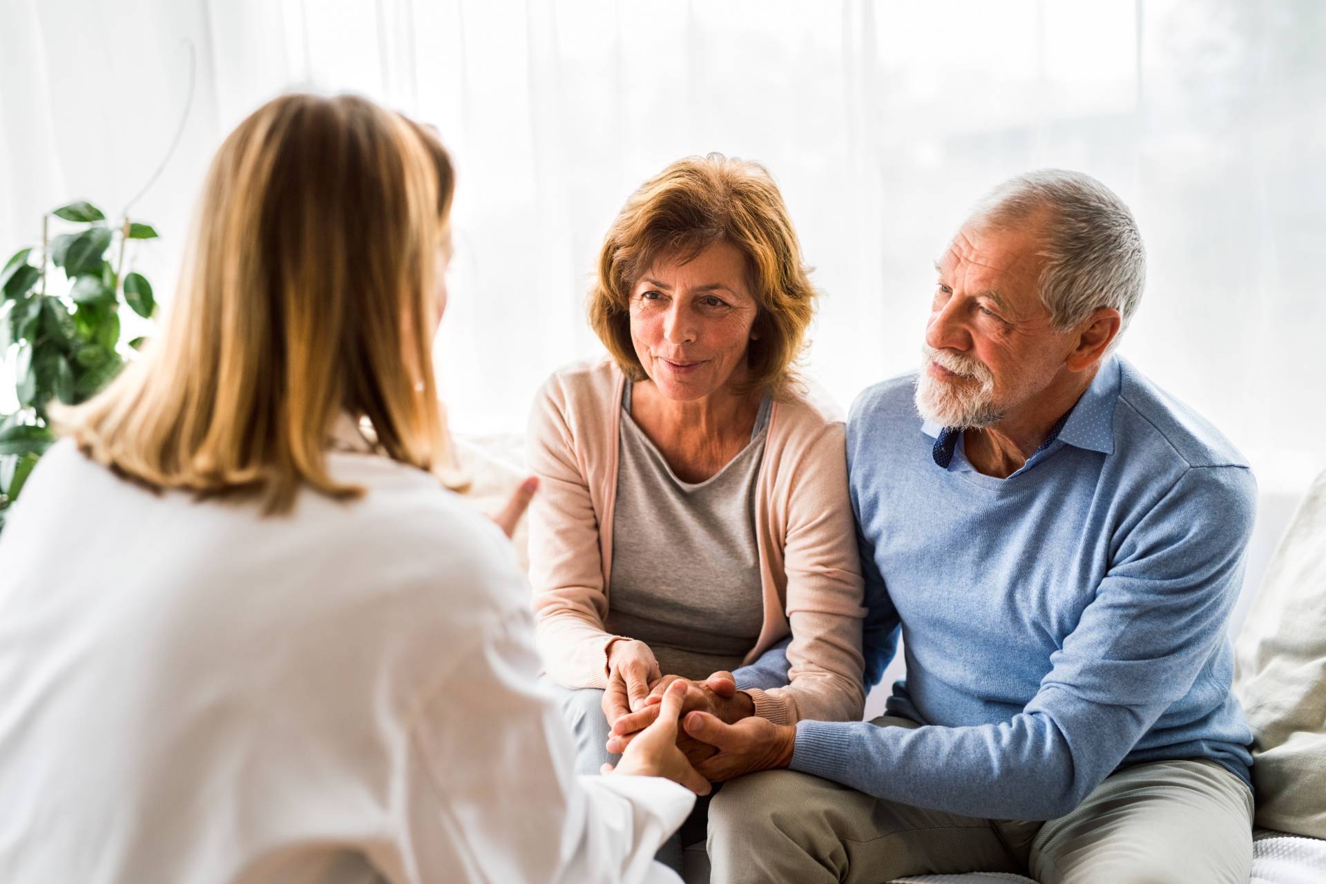 An older couple sitting closely together, holding hands and attentively listening to a professional during a supportive conversation. The setting is bright and comfortable, with soft natural light and a calm atmosphere.